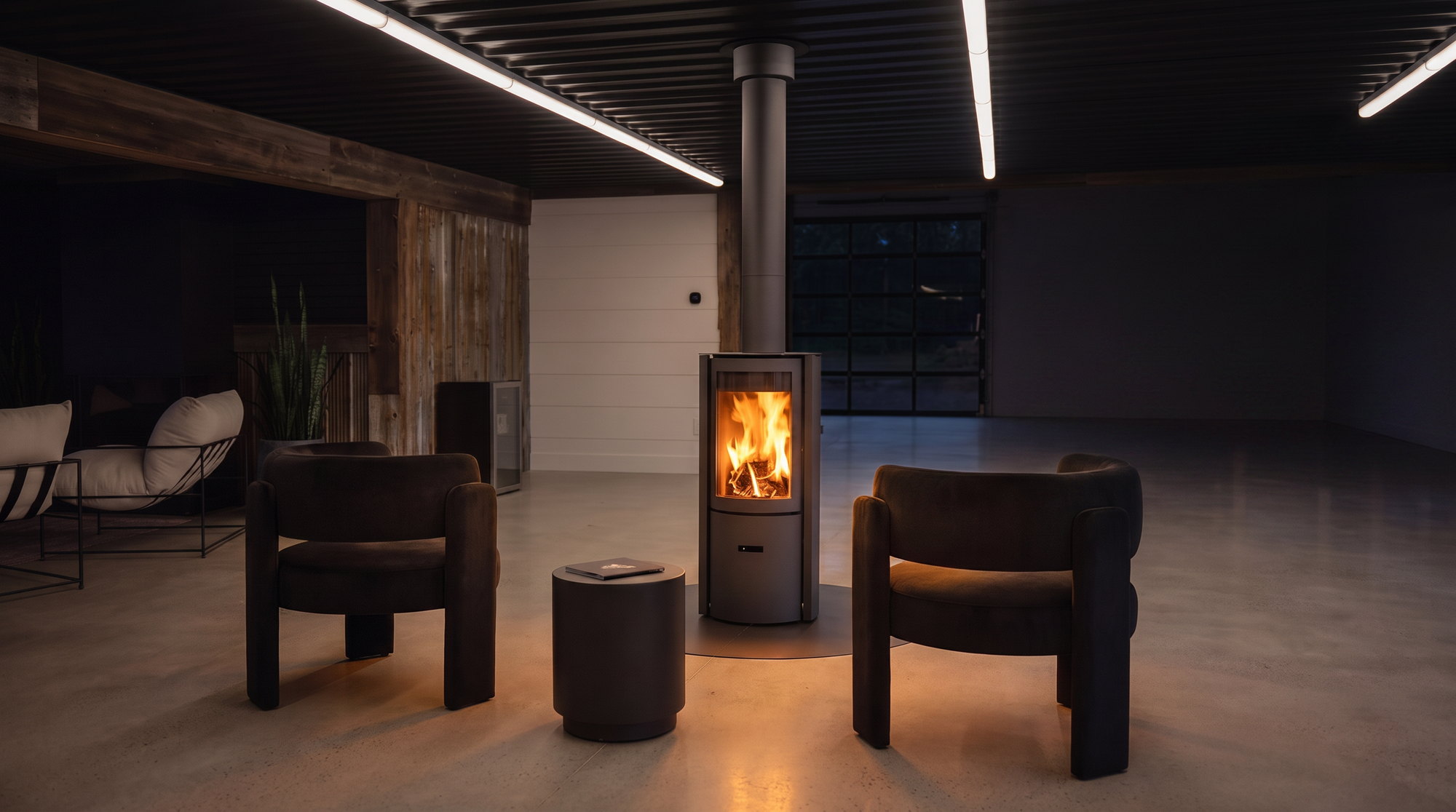 The Stûv 30 stove burning on its round steel floor plate, flanked by two dark velvet chairs under the black corrugated ceiling