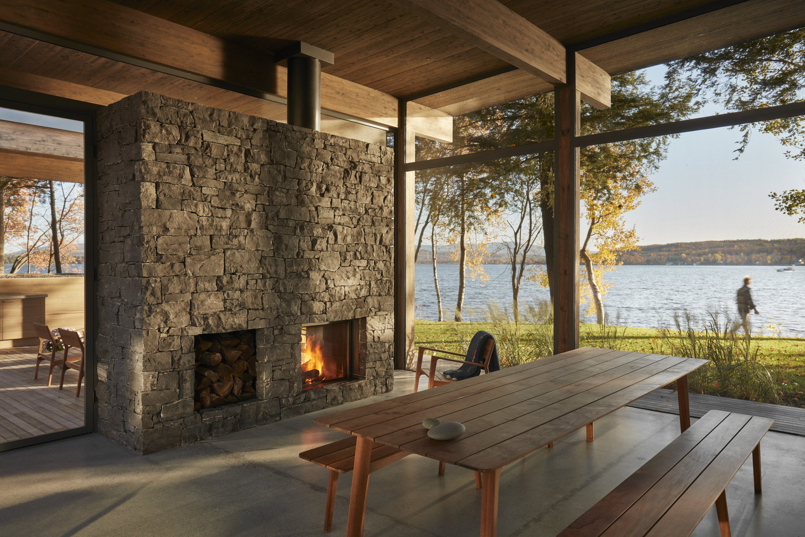 Stûv 21 double-face burning with its glass raised in a great stone chimney on a lakeside porch at Lac-Brome